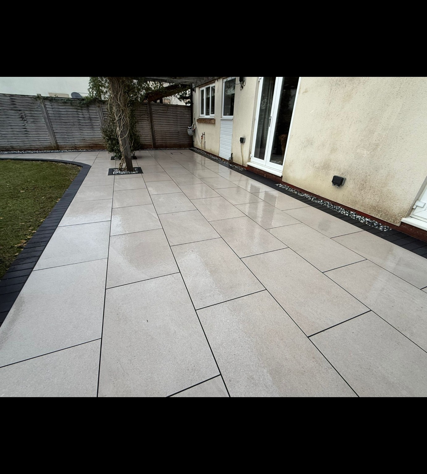Completed porcelain patio in Bridgwater with charcoal edging following a curved lawn and paving fitted around pergola posts